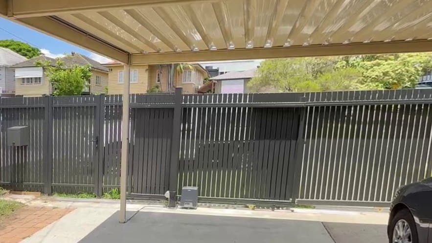 A dark grey aluminium vertical picket sliding gate, closed, seen from under a carport