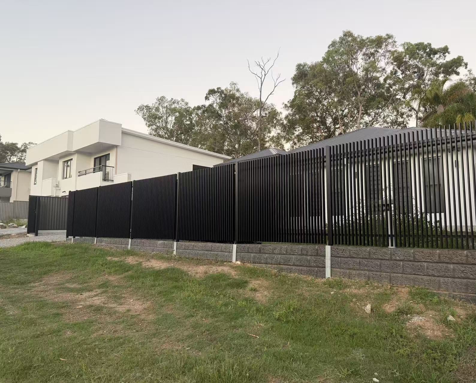 Black aluminium welded vertical blade fence on a split-face block retaining wall above a lawn