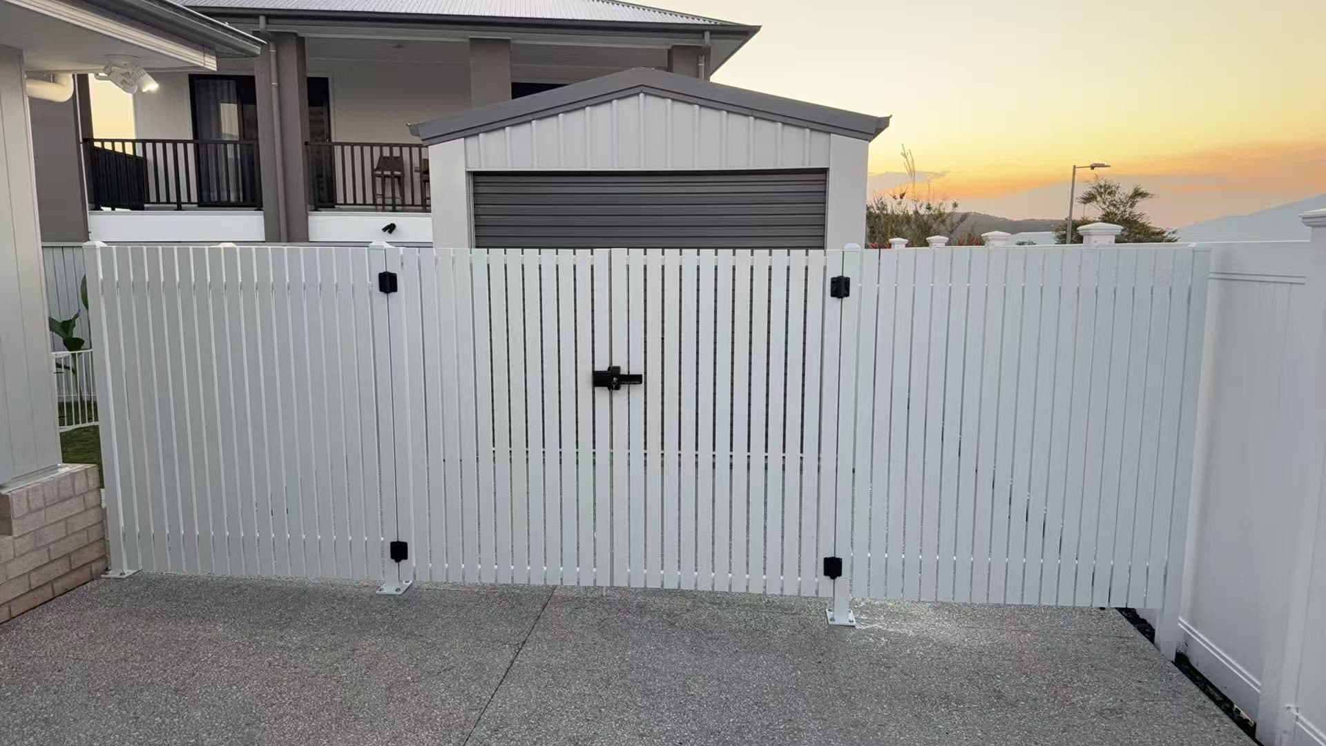 White aluminium vertical picket double swing gate and fence across a concrete driveway in front of a shed