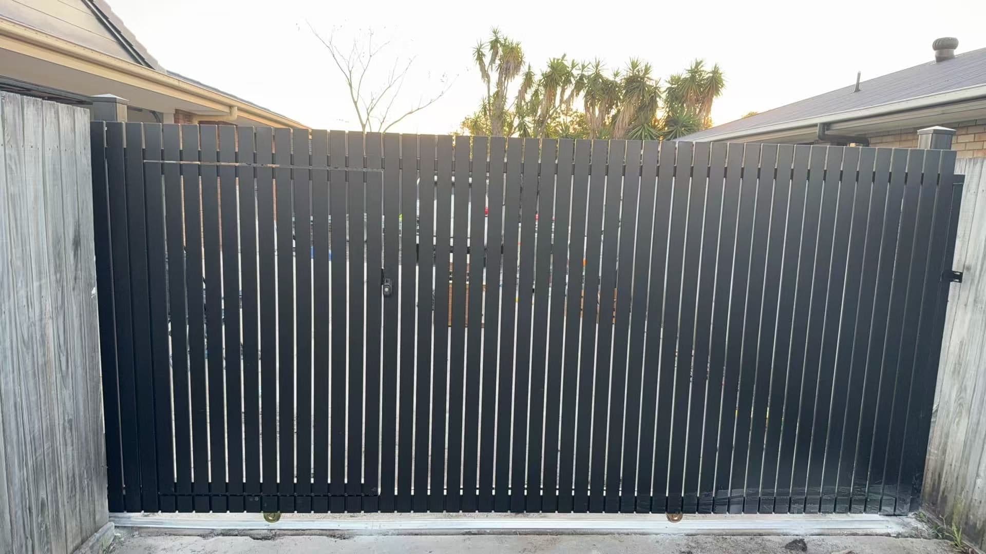 Dark grey aluminium vertical picket sliding driveway gate with a built-in pedestrian door, between timber fences